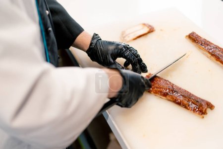 Foto de Primer plano de un chef de sushi con guantes cortando un filete de anguila con cuchillo profesional en la mesa de la cocina mientras preparaba sushi - Imagen libre de derechos
