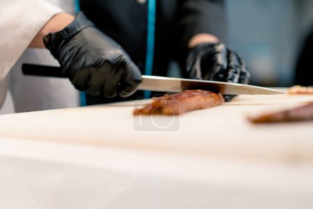 Téléchargez les photos : Gros plan d'un chef sushi en gants coupant un filet d'anguille avec un couteau professionnel sur la table de cuisine tout en faisant des sushis - en image libre de droit