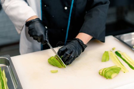 Foto de Primer plano de un fabricante de sushi con guantes cortando aguacate con un cuchillo de cocina profesional en una tabla blanca en la cocina profesional mientras se hace sushi - Imagen libre de derechos