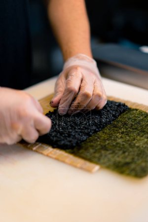 Photo for Close-up of a sushi chef spreading black rice on a nori sheet in the process of making rolls in the kitchen of sushi restaurant - Royalty Free Image