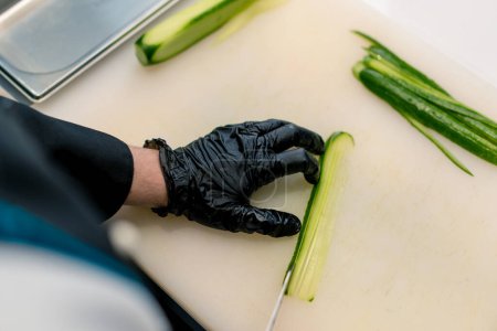 Foto de Primer plano de un fabricante de sushi con guantes cortando un pepino con un cuchillo de cocina profesional en una tabla blanca en una cocina profesional mientras se hace sushi - Imagen libre de derechos