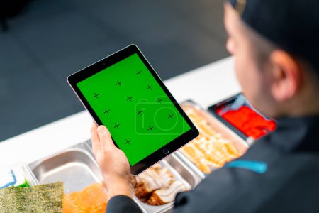 Foto de Primer plano detrás del chef de sushi sosteniendo una tableta con pantalla verde abierta mientras se hace sushi y rollos - Imagen libre de derechos