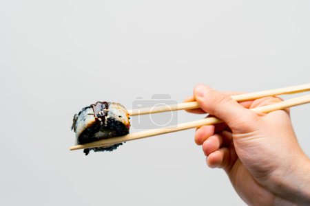 Téléchargez les photos : Gros plan de la californie des sushis avec du riz noir et de l'anguille qu'un homme tient avec des baguettes sur fond blanc - en image libre de droit
