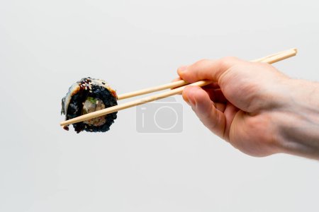 Téléchargez les photos : Gros plan de la californie des sushis avec du riz noir et de l'anguille qu'un homme tient avec des baguettes sur fond blanc - en image libre de droit