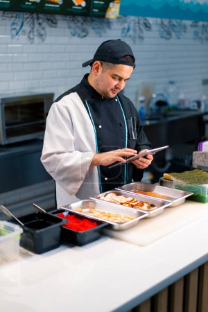 Photo for Thoughtful Sushi Chef with tablet in his hands near the kitchen table on which the products for rolls are laid out tobiko roe salmon shrimp shrimp - Royalty Free Image