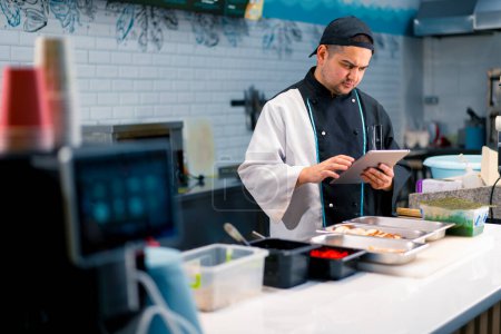 Téléchargez les photos : Chef Sushi réfléchi avec tablette dans ses mains près de la table de cuisine sur laquelle les produits pour rouleaux sont disposés crevettes saumon tobiko oeufs crevettes - en image libre de droit
