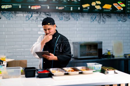 Foto de Chef Sushi reflexivo con la tableta en las manos cerca de la mesa de la cocina en la que se exponen los productos para rollos tobiko roe camarones salmón - Imagen libre de derechos