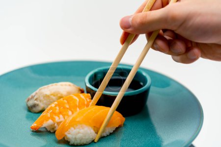 Foto de Primer plano de la mano de un hombre usando palillos para tomar un sushi del plato azul en el que se encuentran tres sushi con camarones de salmón y anguila junto a la salsa de soja - Imagen libre de derechos