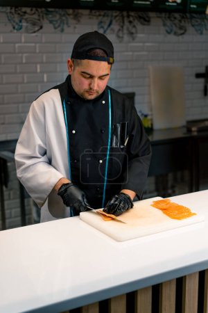 Téléchargez les photos : Gros plan d'un chef sushi coupant un filet de saumon avec un couteau tout en préparant des sushis dans la cuisine du restaurant de sushis - en image libre de droit