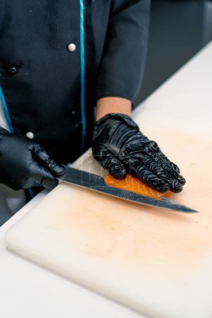 Téléchargez les photos : Gros plan d'un chef sushi coupant un filet de saumon avec un couteau tout en préparant des sushis dans la cuisine du restaurant de sushis - en image libre de droit