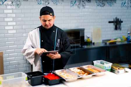 Foto de Chef Sushi reflexivo con la tableta en las manos cerca de la mesa de la cocina en la que se exponen los productos para rollos tobiko roe camarones salmón - Imagen libre de derechos