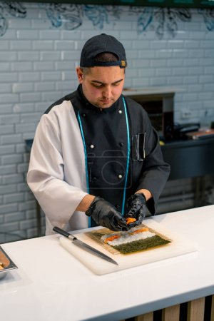 Foto de Primer plano del fabricante de sushi en guantes negros preparando sushi usando hojas de arroz nori salmón filete cuajada queso pepino aguacate en la mesa de la cocina - Imagen libre de derechos