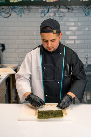 Téléchargez les photos : Gros plan du fabricant de sushis en gants noirs préparant des sushis à l'aide de feuilles de riz nori filet de saumon fromage caillé avocat concombre sur la table de cuisine - en image libre de droit
