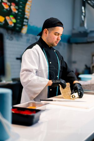 Foto de Chef de sushi en el proceso de preparación de un rollo filipino con queso crema de salmón chamuscado y pepino en una pizarra de cocina blanca - Imagen libre de derechos