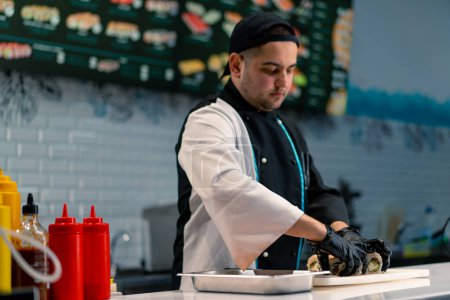 Foto de Primer plano de un hombre de sushi con guantes negros que muestra maravillosamente un rollo de california en un tablero de cocina blanco en la cocina profesional - Imagen libre de derechos