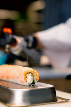 Photo for Close-up of a sushi chef with a gas burner in hand scorching salmon on a Philadelphia roll before serving it at sushi restaurant - Royalty Free Image