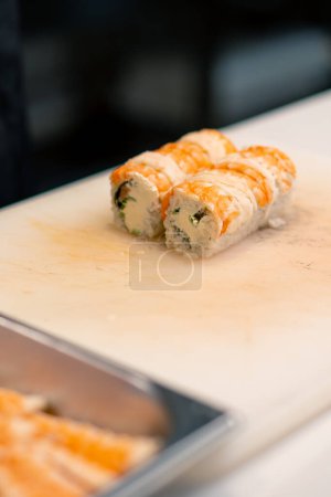 Téléchargez les photos : Gros plan d'un homme de sushi en gants noirs coupant un rouleau de californie sur un tableau de cuisine blanc dans une cuisine professionnelle - en image libre de droit