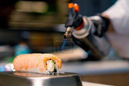 Foto de Primer plano de un chef de sushi con un quemador de gas en la mano salmón abrasador en un rollo de Filadelfia antes de servirlo en el restaurante de sushi - Imagen libre de derechos