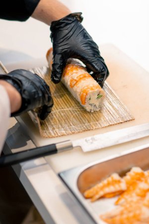 Foto de Primer plano de un fabricante de sushi con guantes negros envolviendo un rollo de california con una estera de sushi de bambú en la cocina profesional - Imagen libre de derechos