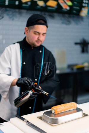 Photo for Close-up of a sushi chef with a gas burner in hand scorching salmon on a Philadelphia roll before serving it at sushi restaurant - Royalty Free Image