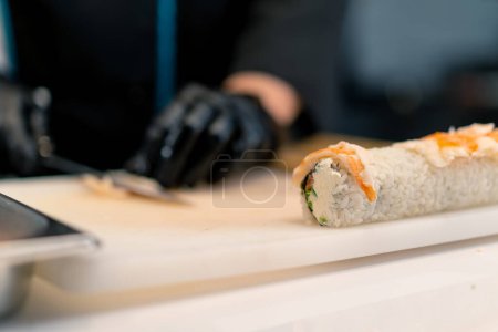 Téléchargez les photos : Gros plan d'un fabricant de sushis en gants noirs coupant un filet de crevettes avec un couteau tout en préparant un rouleau de californie dans la cuisine - en image libre de droit