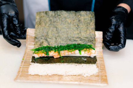 Foto de Chef de sushi en el proceso de preparación de rollo de Filadelfia con camarones de queso crema de salmón y algas chuka en una estera de sushi de bambú - Imagen libre de derechos