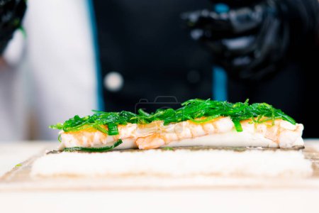 Téléchargez les photos : Chef sushi en train de préparer un petit pain Philadelphie avec des crevettes au fromage à la crème de saumon et des algues chuka sur un tapis de sushi en bambou - en image libre de droit