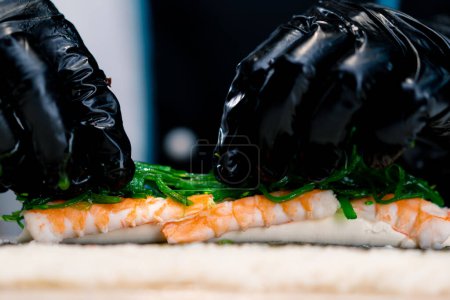 Téléchargez les photos : Chef sushi en train de préparer un petit pain Philadelphie avec des crevettes au fromage à la crème de saumon et des algues chuka sur un tapis de sushi en bambou - en image libre de droit