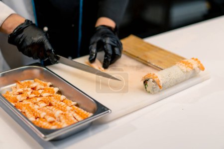 Photo for Close-up of sushi maker in black gloves preparing sushi using rice nori sheets salmon fillet curd cheese cucumber avocado on the kitchen table - Royalty Free Image
