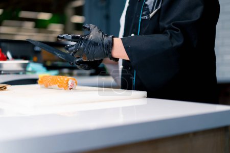 Foto de Chef de sushi corta un rollo de Filadelfia que cocinó con queso crema de salmón y pepino con un cuchillo en la cocina del restaurante de sushi - Imagen libre de derechos