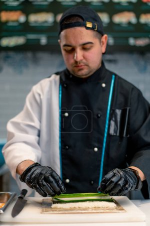 Téléchargez les photos : Gros plan du chef sushi en gants noirs préparant des sushis à l'aide de feuilles de riz nori filet de crevettes crevettes caillé fromage concombre avocat sur la table de cuisine - en image libre de droit