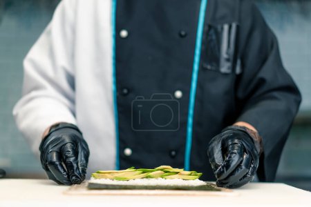 Foto de Un chef de sushi prepara rollos de maki con arroz nori de aguacate y una esterilla de bambú para hacer sushi en la cocina del restaurante de sushi. - Imagen libre de derechos