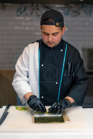 Téléchargez les photos : Un chef de sushi prépare des rouleaux de maki en utilisant du riz à l'avocat nori et un tapis de bambou pour faire des sushis dans la cuisine du restaurant de sushi - en image libre de droit