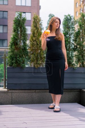 Téléchargez les photos : Portrait d'une jeune fille souriante avec un cocktail dans les mains debout sur une terrasse d'été dans un bar-club - en image libre de droit