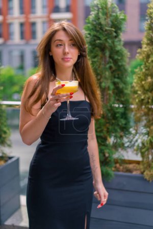 Photo for Portrait of a young smiling girl with a cocktail in her hands standing on a summer terrace in bar-club - Royalty Free Image
