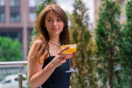 Téléchargez les photos : Portrait d'une jeune fille souriante avec un cocktail dans les mains debout sur une terrasse d'été dans un bar-club - en image libre de droit