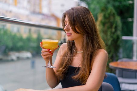 Téléchargez les photos : Une jeune fille souriante avec un cocktail dans les mains et de le boire et assis à une table sur une terrasse d'été surplombant la ville dans le bar club - en image libre de droit