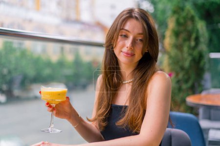 Foto de Una joven sonriente con un cóctel en sus manos se sienta en una mesa en una terraza de verano con vistas a la ciudad en el bar club - Imagen libre de derechos
