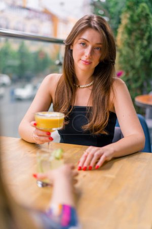 Foto de Una joven sonriente con un cóctel en sus manos se sienta y charla con un amigo en una mesa en la terraza de verano con vistas a la ciudad en el bar club - Imagen libre de derechos