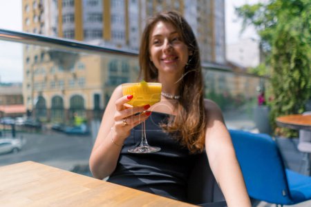 Photo for A young smiling girl with a cocktail in her hands sits at a table on a summer terrace overlooking the city in bar club - Royalty Free Image