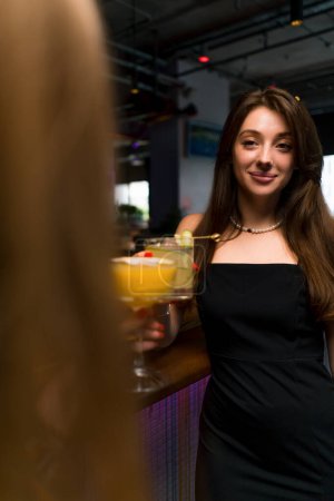Foto de Una joven sonriente con un cóctel en sus manos está de pie en el mostrador del bar y hablando con su amigo en el bar club - Imagen libre de derechos