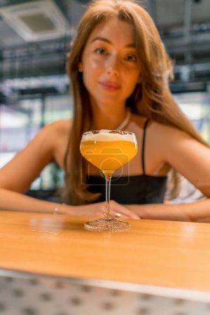 Téléchargez les photos : Portrait d'une jeune fille souriante derrière un bar dans un club avec un délicieux cocktail alcoolisé - en image libre de droit