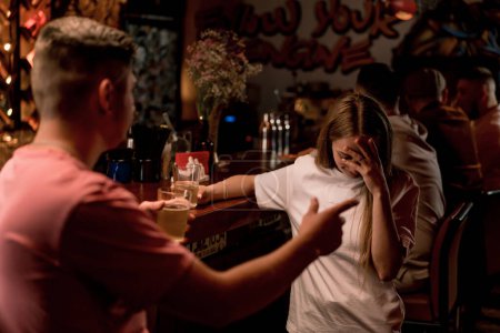 Photo for An upset guy with a mustache stands with a glass of beer in his hand at the bar and argues with girl in a club bar - Royalty Free Image