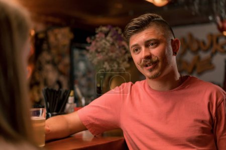 Photo for A cheerful guy with a mustache is standing with a glass of beer in his hand at the bar and chatting with attractive girl in a club bar - Royalty Free Image