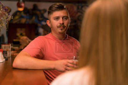Foto de Un chico alegre con bigote está de pie con un vaso de cerveza en la mano en el bar y charlando con una chica atractiva en un bar del club - Imagen libre de derechos
