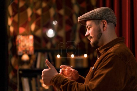 Téléchargez les photos : Un homme souriant dans une casquette et une chemise tenant un verre avec un cocktail au bar du club - en image libre de droit