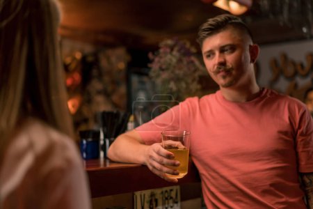Téléchargez les photos : Un gars joyeux avec une moustache est debout avec un verre de bière dans la main au bar et bavarder avec une jolie fille dans un bar club - en image libre de droit
