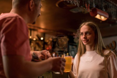 Téléchargez les photos : Une jeune fille souriante avec un verre de bière à la main discute avec un homme au bar du club bar - en image libre de droit
