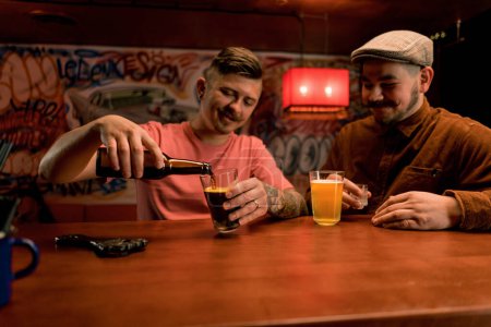 Photo for Two friends of a friend are sitting at the bar having fun chatting and pouring beer into glasses in club bar - Royalty Free Image