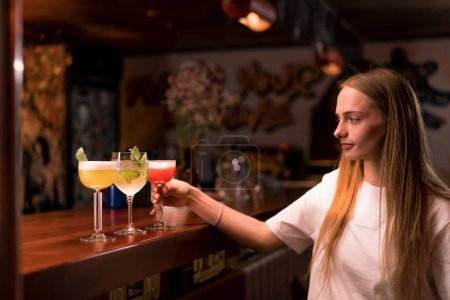 Photo for A young girl takes one of the cocktails from the bar counter in club bar concept of love for alcoholic beverages and their mixing - Royalty Free Image